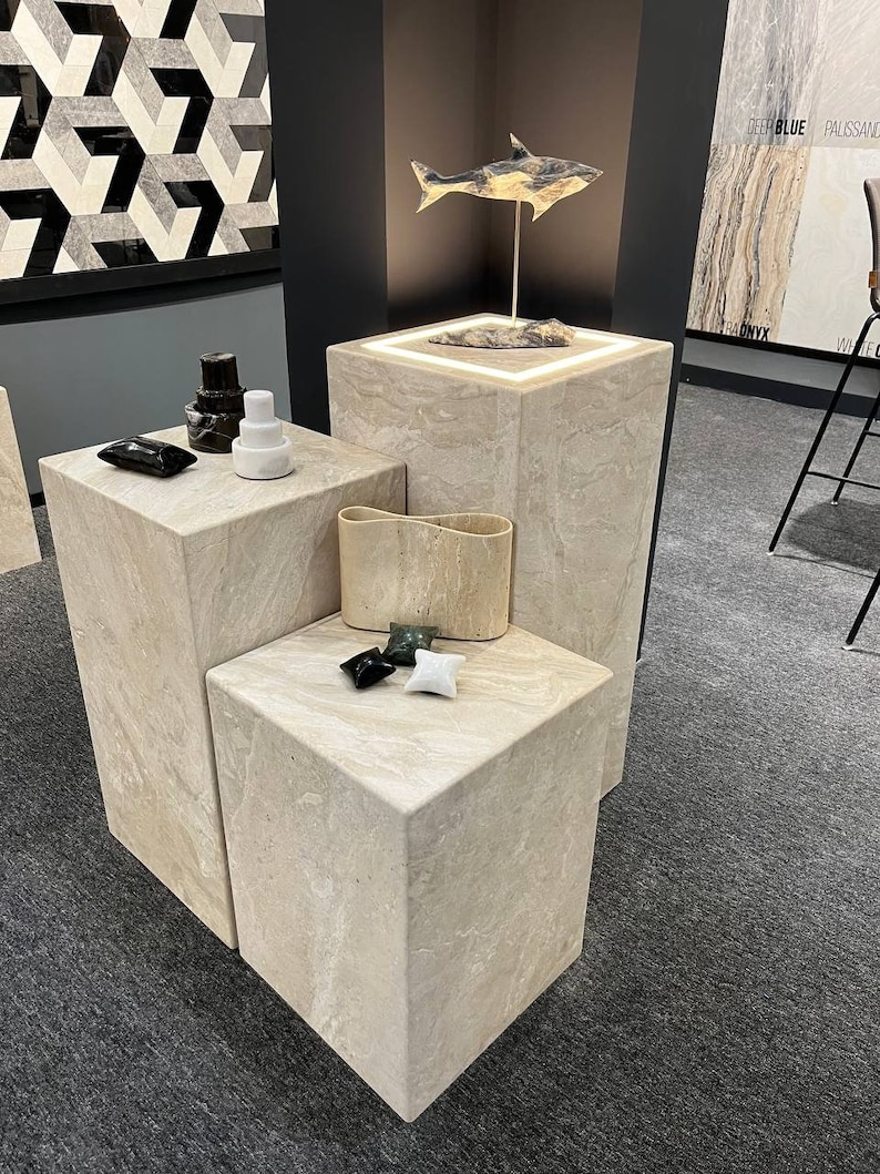 Puede incluir: Tres expositores de piedra beige con una escultura de pez de madera en el expositor superior. Los expositores est&aacute;n sobre una alfombra gris.