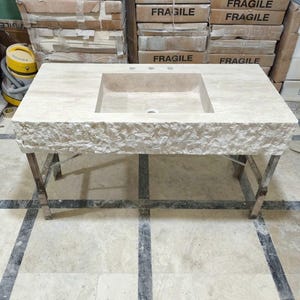 May include: A rectangular beige stone vanity top featuring a central integrated sink basin with three faucet holes. The front edge of the vanity displays a rough, chiseled natural stone texture, while the top surface is smooth and polished.