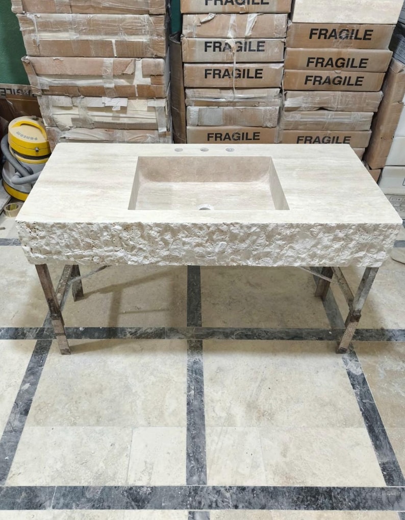 May include: A rectangular beige stone vanity top featuring a central integrated sink basin with three faucet holes. The front edge of the vanity displays a rough, chiseled natural stone texture, while the top surface is smooth and polished.