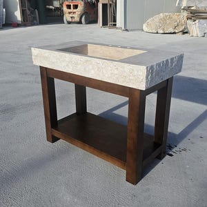 May include: A bathroom vanity featuring a rectangular, light beige natural stone sink basin with a rough, chiseled edge. The sink is supported by a dark brown wooden base with four straight legs and a lower shelf for storage and organization.