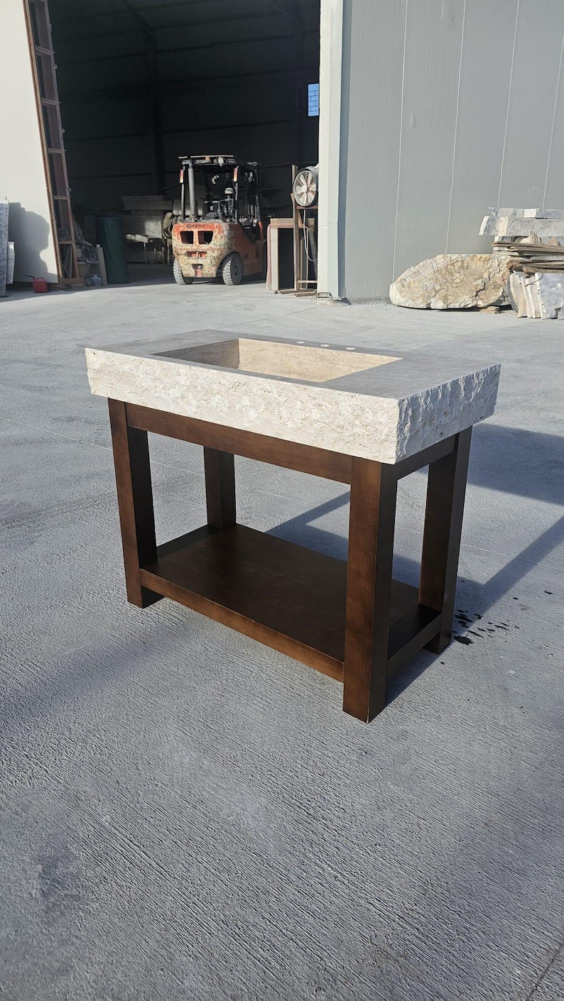 May include: A bathroom vanity featuring a rectangular, light beige natural stone sink basin with a rough, chiseled edge. The sink is supported by a dark brown wooden base with four straight legs and a lower shelf for storage and organization.