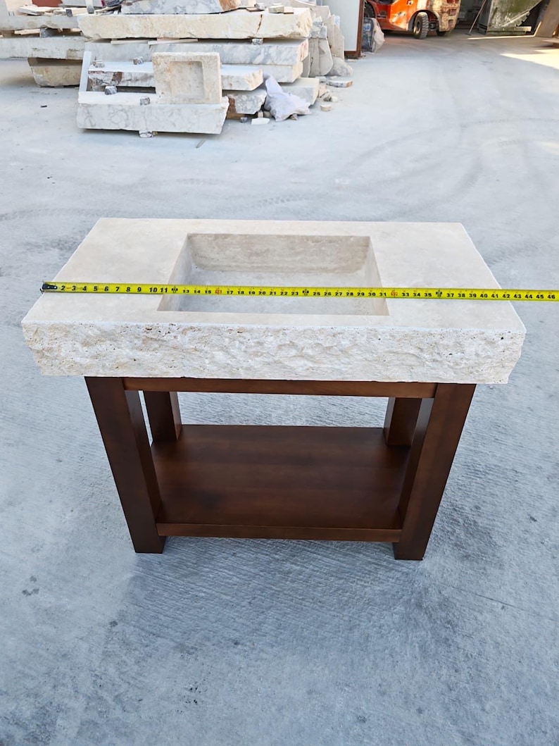 May include: A rectangular beige stone sink with a rough-hewn front edge, mounted on a dark brown wooden vanity base with a lower shelf. A yellow measuring tape is stretched across the top, showing a width of approximately 45 inches.