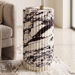 May include: A cylindrical side table with a fluted design. The table is made of white marble with dark gray veining. The table is approximately 20 inches tall and has a gold accent at the top.