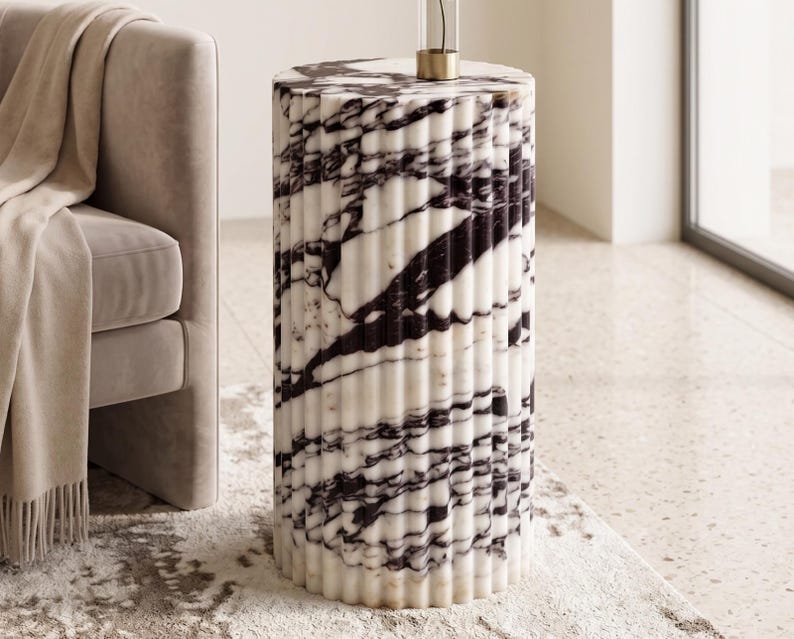 May include: A cylindrical side table with a fluted design. The table is made of white marble with dark gray veining. The table is approximately 20 inches tall and has a gold accent at the top.