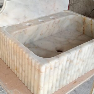 May include: A white marble sink with a fluted edge and three holes for faucets. The sink is rectangular and has a drain in the bottom.