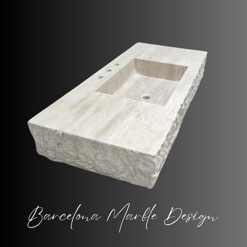 May include: A rectangular beige stone bathroom sink with a smooth top surface and a rough, chiseled natural edge. The basin features a central drain hole and three pre-drilled faucet holes on the deck. The text Barcelona Marble Design is below.