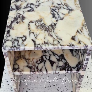 K&ouml;nnte beinhalten: Ein wei&szlig;er Marmor-Beistelltisch mit einem zweistufigen Design. Der Tisch hat eine wei&szlig;e Marmorplatte mit schwarzen und grauen Adern. Die untere Stufe ist ebenfalls aus wei&szlig;em Marmor mit schwarzen und grauen Adern. Der Tisch steht auf einem wei&szlig;en Fliesenboden. Barcelona Marble Designs ist auf der Vorderseite des Tisches geschrieben.