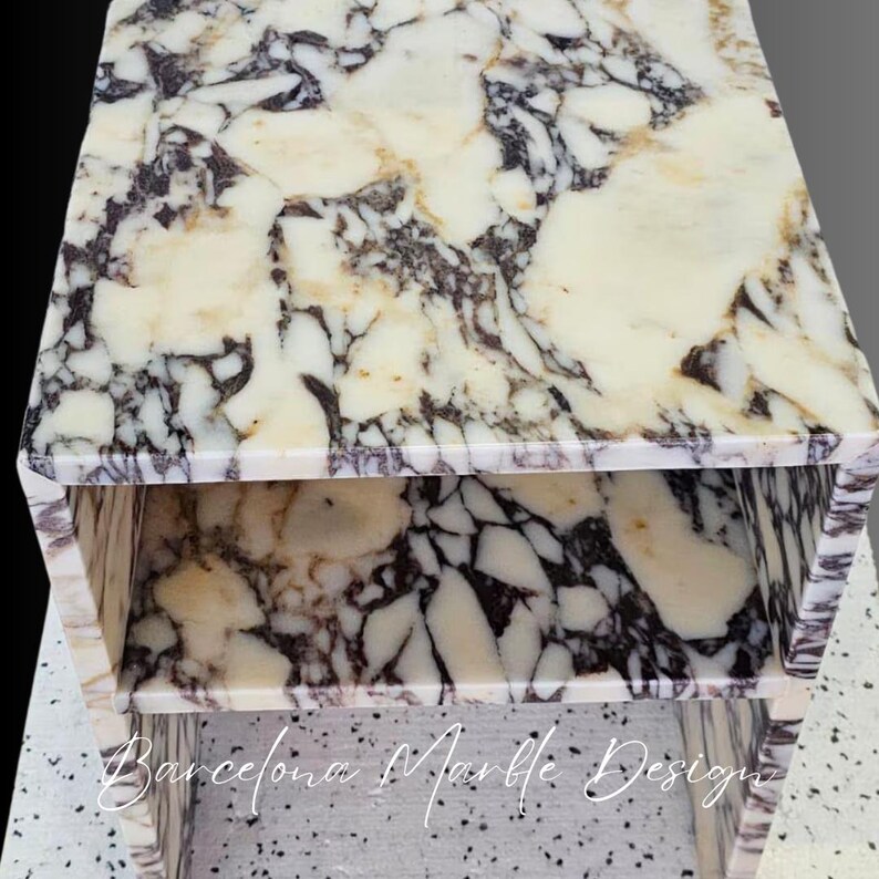 K&ouml;nnte beinhalten: Ein wei&szlig;er Marmor-Beistelltisch mit einem zweistufigen Design. Der Tisch hat eine wei&szlig;e Marmorplatte mit schwarzen und grauen Adern. Die untere Stufe ist ebenfalls aus wei&szlig;em Marmor mit schwarzen und grauen Adern. Der Tisch steht auf einem wei&szlig;en Fliesenboden. Barcelona Marble Designs ist auf der Vorderseite des Tisches geschrieben.