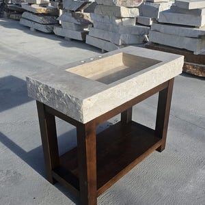 May include: A rectangular bathroom vanity featuring a beige, porous stone countertop with a chiseled, rough-hewn edge and a rectangular basin. The countertop rests on a dark brown wooden base with four straight legs and a lower storage shelf.