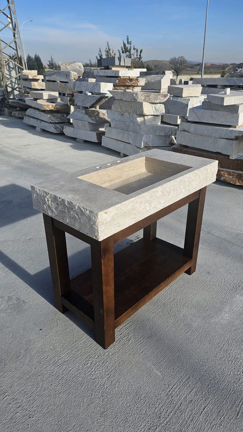 May include: A rectangular bathroom vanity featuring a beige, porous stone countertop with a chiseled, rough-hewn edge and a rectangular basin. The countertop rests on a dark brown wooden base with four straight legs and a lower storage shelf.
