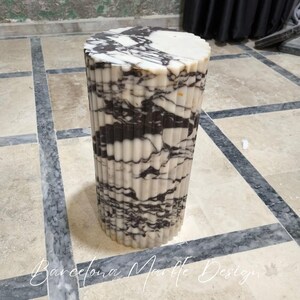 May include: A cylindrical marble side table with a fluted design. The table features a white and black marbled pattern. The top is a solid white color. The words "Barcelona Marble Design" are at the bottom of the image.