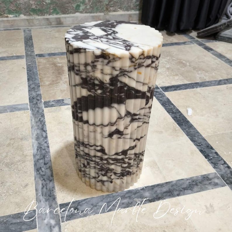 May include: A cylindrical marble side table with a fluted design. The table features a white and black marbled pattern. The top is a solid white color. The words "Barcelona Marble Design" are at the bottom of the image.