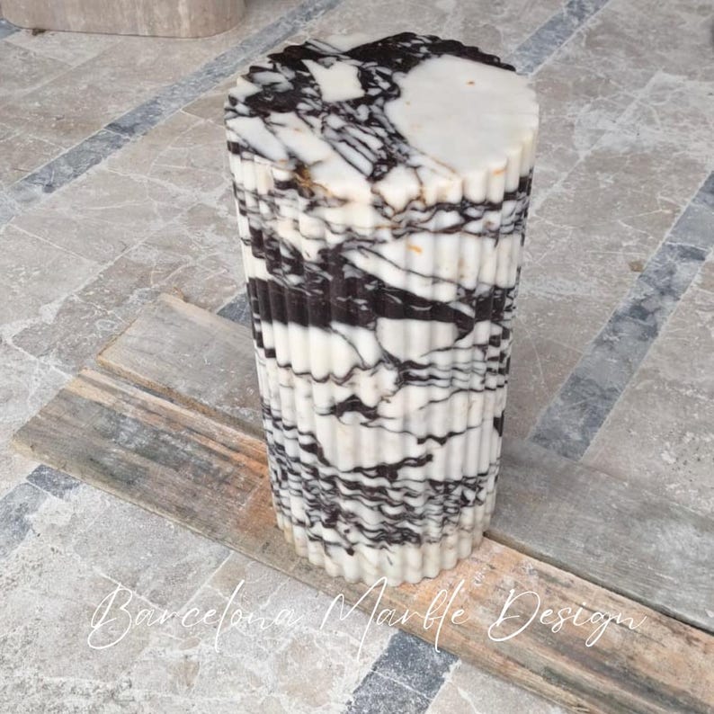 May include: A cylindrical marble pedestal with a fluted design. The stone features a striking pattern of black and white veining. The top is a solid white color. The pedestal is a decorative element.