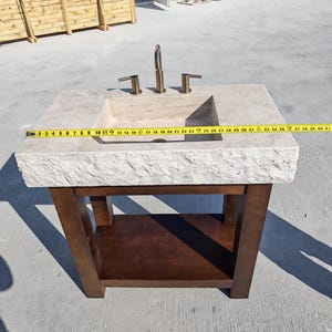 May include: A beige stone vanity sink with a rough-hewn front edge and a dark brown wooden base. A yellow measuring tape is stretched across the top, showing a width of approximately 40 inches. A brass-colored faucet is mounted on the stone surface.