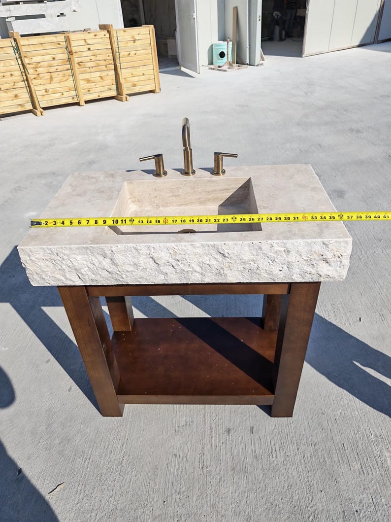 May include: A beige stone vanity sink with a rough-hewn front edge and a dark brown wooden base. A yellow measuring tape is stretched across the top, showing a width of approximately 40 inches. A brass-colored faucet is mounted on the stone surface.