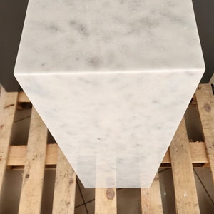 May include: White marble pedestal with a smooth, polished surface. The pedestal is rectangular and sits on a wooden pallet.