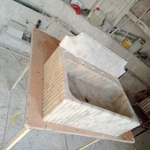 May include: A white marble sink with a decorative, fluted design. The sink is mounted on a wooden table.