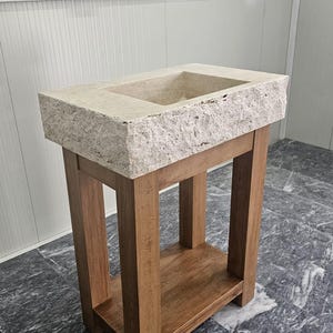 May include: A rectangular beige stone sink with a rough, textured exterior edge and a smooth basin, mounted on a brown wooden stand featuring four vertical legs and a lower shelf for storage, providing a sturdy base for the heavy stone basin.