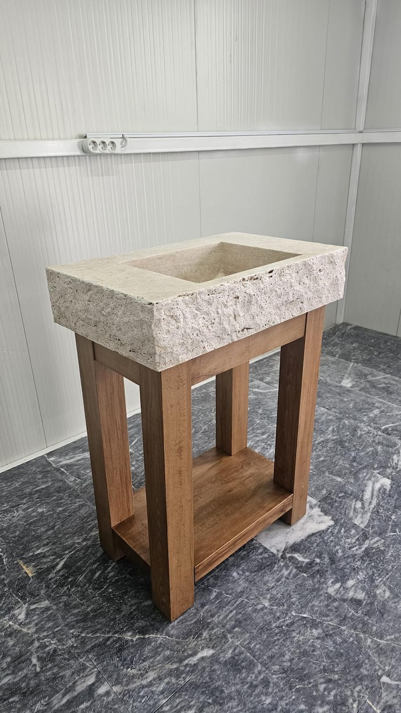 May include: A rectangular beige stone sink with a rough, textured exterior edge and a smooth basin, mounted on a brown wooden stand featuring four vertical legs and a lower shelf for storage, providing a sturdy base for the heavy stone basin.
