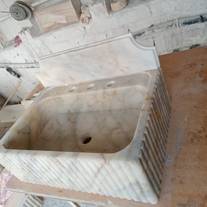 May include: A white marble bathroom sink with a decorative, fluted base and a backsplash. The sink has three holes for faucets.