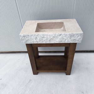 May include: A rectangular stone sink with a rough, chiseled front edge and a smooth top surface, mounted on a dark brown wooden vanity base. The vanity features four sturdy legs and a lower shelf, providing an open storage area beneath the basin.