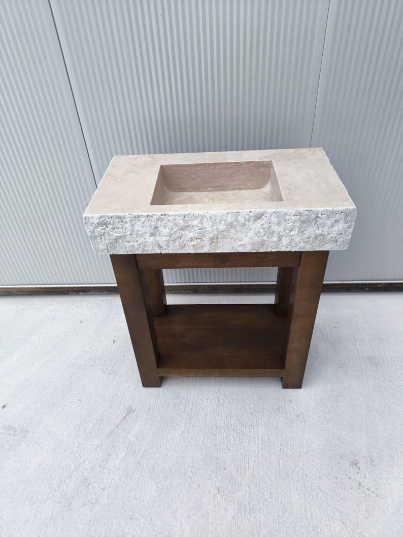 May include: A rectangular stone sink with a rough, chiseled front edge and a smooth top surface, mounted on a dark brown wooden vanity base. The vanity features four sturdy legs and a lower shelf, providing an open storage area beneath the basin.