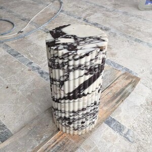 May include: A cylindrical marble pedestal with a fluted design. The top surface is round and features black and white veining. The body of the pedestal has vertical fluting with alternating bands of black and white. It is sitting on a wooden plank.