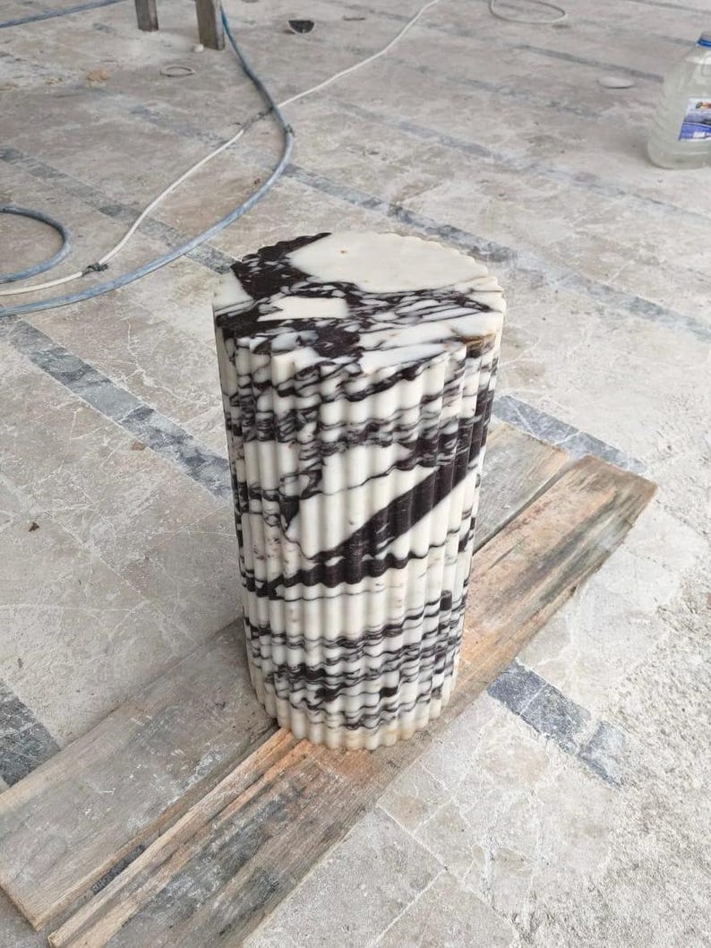 May include: A cylindrical marble pedestal with a fluted design. The top surface is round and features black and white veining. The body of the pedestal has vertical fluting with alternating bands of black and white. It is sitting on a wooden plank.