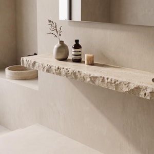 May include: A bathroom interior with a stone shelf holding a vase, a dark brown bottle, and a candle. A folded towel is on the shelf. A round bowl sits below. The room has a minimalist design with neutral tones.