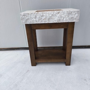 May include: A rectangular bathroom vanity featuring a thick, light-colored stone sink basin with a rough, textured exterior edge. The sink is supported by a dark brown wooden base with four sturdy legs and a lower shelf for storage.