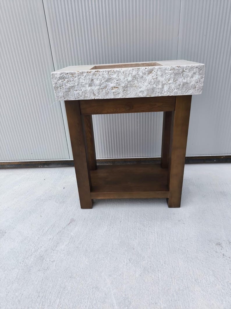 May include: A rectangular bathroom vanity featuring a thick, light-colored stone sink basin with a rough, textured exterior edge. The sink is supported by a dark brown wooden base with four sturdy legs and a lower shelf for storage.