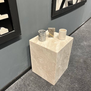 Puede incluir: Un peque&ntilde;o pedestal cuadrado de m&aacute;rmol beige claro con tres objetos encima. Los objetos son un peque&ntilde;o recipiente cil&iacute;ndrico gris, un peque&ntilde;o objeto rectangular marr&oacute;n y un peque&ntilde;o recipiente cil&iacute;ndrico blanco.