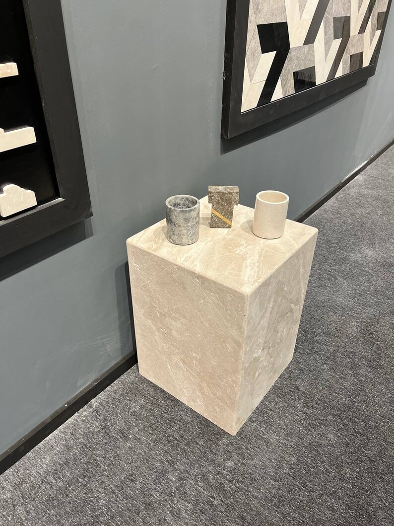 Puede incluir: Un peque&ntilde;o pedestal cuadrado de m&aacute;rmol beige claro con tres objetos encima. Los objetos son un peque&ntilde;o recipiente cil&iacute;ndrico gris, un peque&ntilde;o objeto rectangular marr&oacute;n y un peque&ntilde;o recipiente cil&iacute;ndrico blanco.