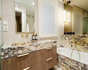 May include: A modern bathroom featuring a marble countertop and vanity with two drawers. The vanity has gold hardware and a large arched mirror above it. A white bathtub is on the right side of the image, with a marble surround. The bathroom has a white tiled floor and a white ceiling.