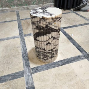 May include: A cylindrical marble side table with a fluted design. The table features a mix of white and black veining, creating a striking pattern. The top is a solid white, and the base is a repeating pattern of vertical lines. The table is approximately 18 inches tall.
