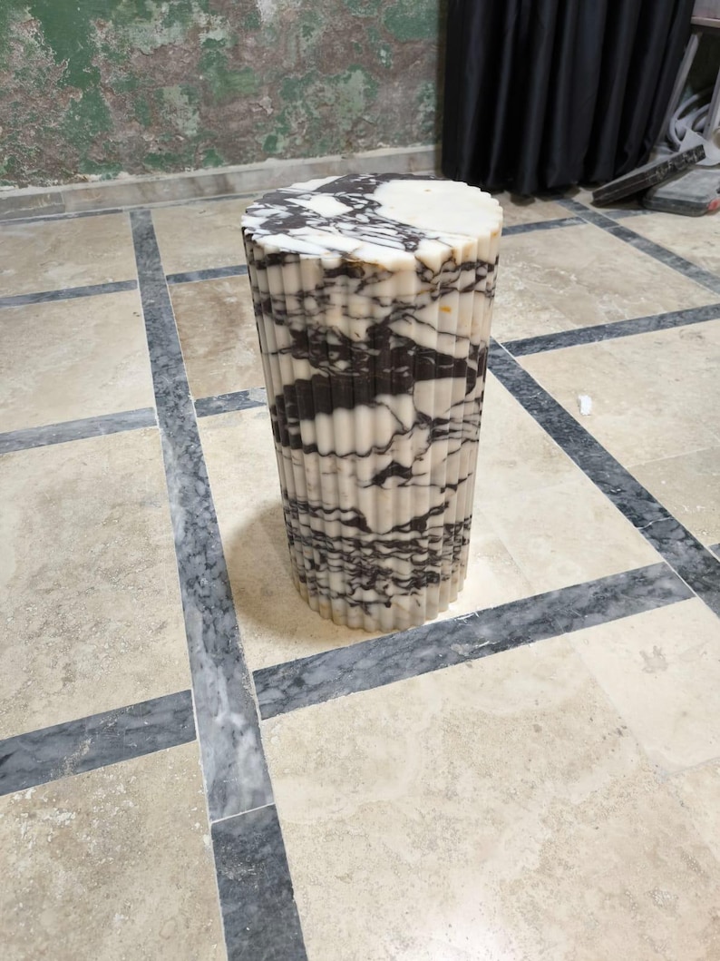 May include: A cylindrical marble side table with a fluted design. The table features a mix of white and black veining, creating a striking pattern. The top is a solid white, and the base is a repeating pattern of vertical lines. The table is approximately 18 inches tall.
