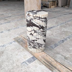May include: A cylindrical marble pillar with a fluted design. The pillar is white with striking black veining. It stands on a wooden plank, showcasing its texture and pattern. The pillar is a decorative element, likely for interior design.