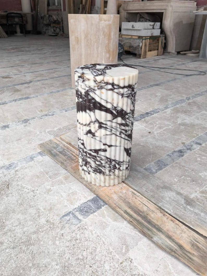 May include: A cylindrical marble pillar with a fluted design. The pillar is white with striking black veining. It stands on a wooden plank, showcasing its texture and pattern. The pillar is a decorative element, likely for interior design.
