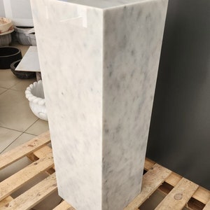 May include: A white marble pedestal with a square top and a rectangular base. The pedestal is sitting on a wooden pallet.