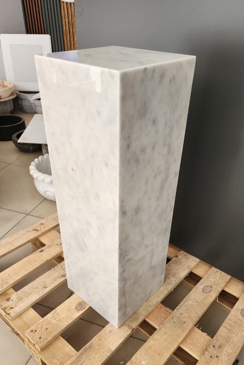May include: A white marble pedestal with a square top and a rectangular base. The pedestal is sitting on a wooden pallet.