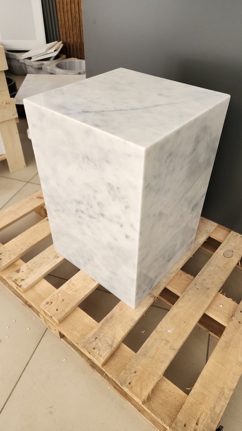 May include: A white marble cube stool with a light gray veining pattern. The stool is sitting on a wooden pallet.