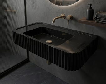 May include: A black marble bathroom sink with a rectangular basin and a fluted, scalloped edge. The sink has a gold-colored drain and faucet. A black soap dispenser and folded towels sit on a wooden shelf above the sink.