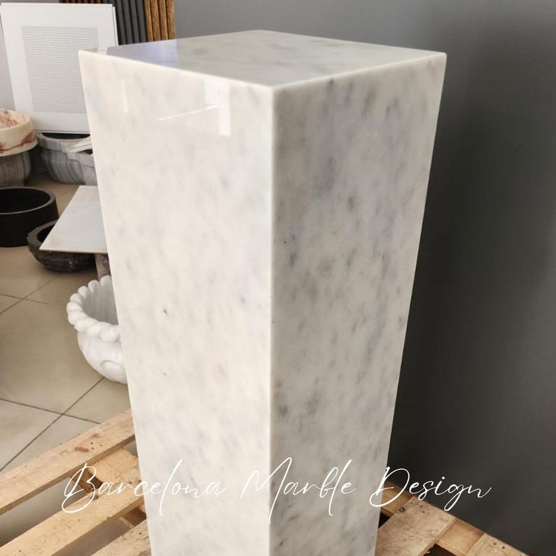 May include: White marble pedestal with a smooth, polished surface. The pedestal is square and has a modern design.
