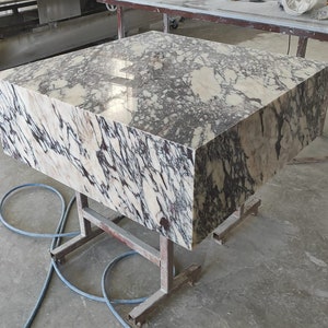 K&ouml;nnte beinhalten: Eine gro&szlig;e, rechteckige Platte aus poliertem Marmor mit einem wei&szlig;en und violetten Adernmuster. Die Platte steht auf einem Metallgestell in einer Werkstatt.