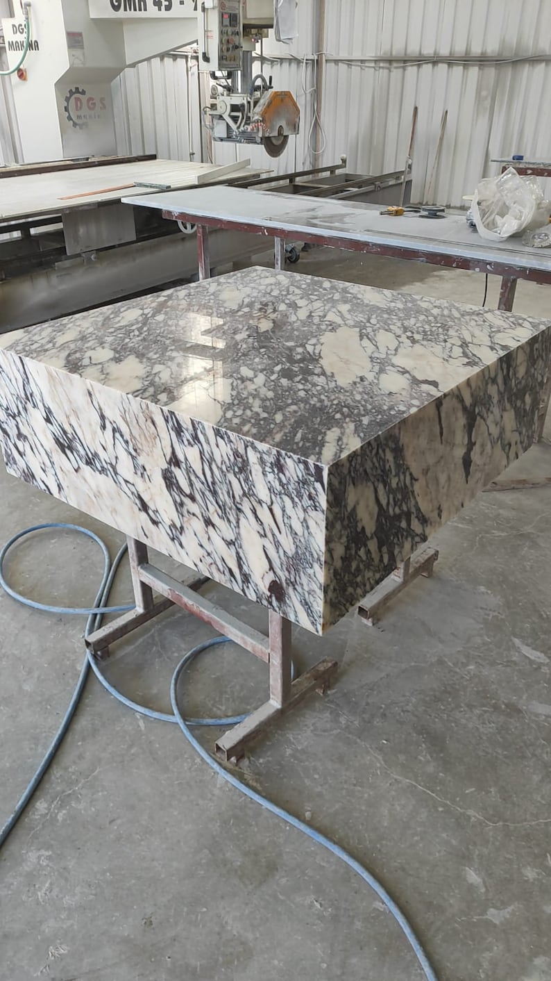 K&ouml;nnte beinhalten: Eine gro&szlig;e, rechteckige Platte aus poliertem Marmor mit einem wei&szlig;en und violetten Adernmuster. Die Platte steht auf einem Metallgestell in einer Werkstatt.