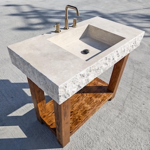 May include: A rectangular stone vanity sink with a rough, chiseled edge and a smooth basin surface. It features a metallic gold-toned faucet with two handles and is supported by a sturdy wooden base with four legs and a lower storage shelf.