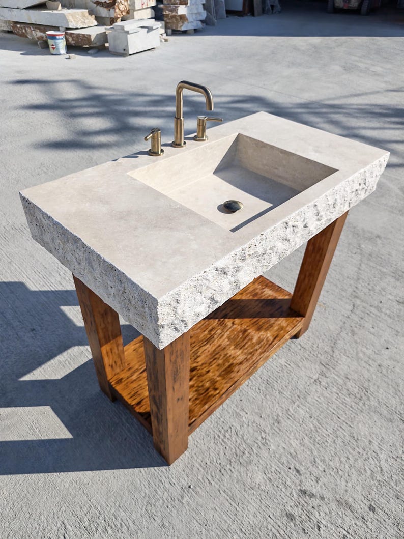 May include: A rectangular stone vanity sink with a rough, chiseled edge and a smooth basin surface. It features a metallic gold-toned faucet with two handles and is supported by a sturdy wooden base with four legs and a lower storage shelf.