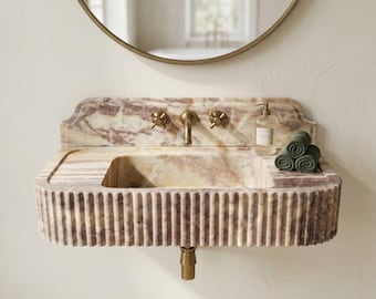 Puede incluir: Un lavabo de baño de mármol en beige y burdeos, con un diseño acanalado y grifos de latón. El lavabo tiene un dispensador de jabón y toallas verdes enrolladas. Un espejo redondo cuelga encima. El diseño recuerda al Art Déco.