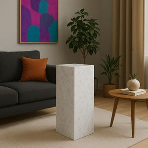 May include: White marble pedestal with a square top and a smooth, polished finish. The pedestal is standing on a light beige rug in a living room setting.