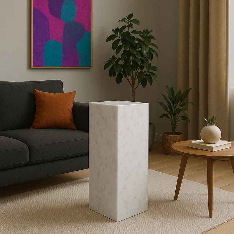 May include: White marble pedestal with a square top and a smooth, polished finish. The pedestal is standing on a light beige rug in a living room setting.